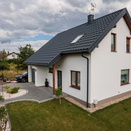 Hygge Hus Sp. z o.o. - Nowoczesny, biały dom z ciemną, skośną dachówką, widok z góry na ogród z kostką brukową i zaparkowanym samochodem, otoczony zielenią i innymi domami w oddali.
