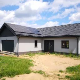 Nowoczesny parterowy dom z szarą elewacją, ciemną dachówką i panelami słonecznymi na dachu, otoczony piaskiem i trawą, błękitne niebo z chmurami w tle.