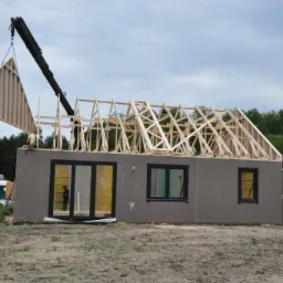 Montaż domu szkieletowego: dźwig podnosi drewniany element konstrukcyjny dachu. Widoczne ściany z oknami i częściowo ukończona więźba dachowa. Etap budowy.