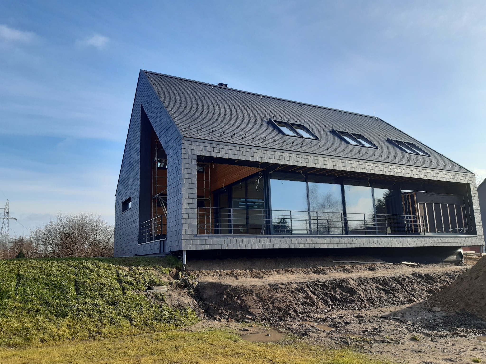 Nowoczesny dom z szarej dachówki, duże okna z metalową balustradą, teren wokół domu w trakcie prac ziemnych, widoczne ślady błota.