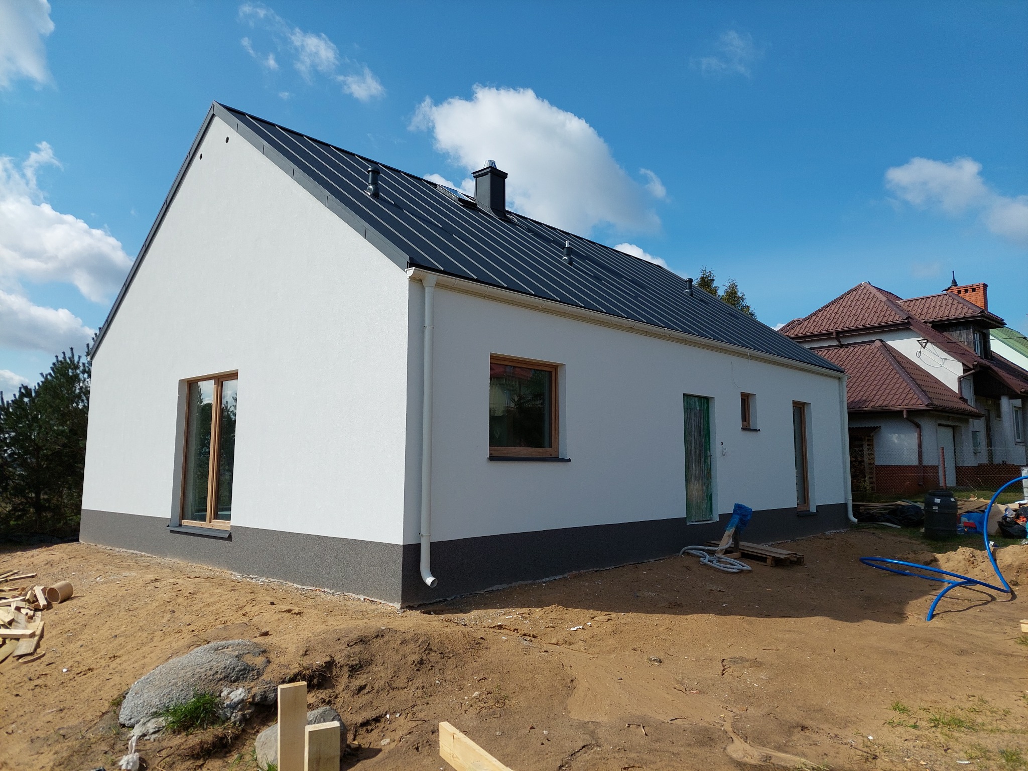 Nowoczesny dom z białymi ścianami i grafitowym dachem z blachy na działce w trakcie zagospodarowania, widoczne elementy instalacji zewnętrznej.