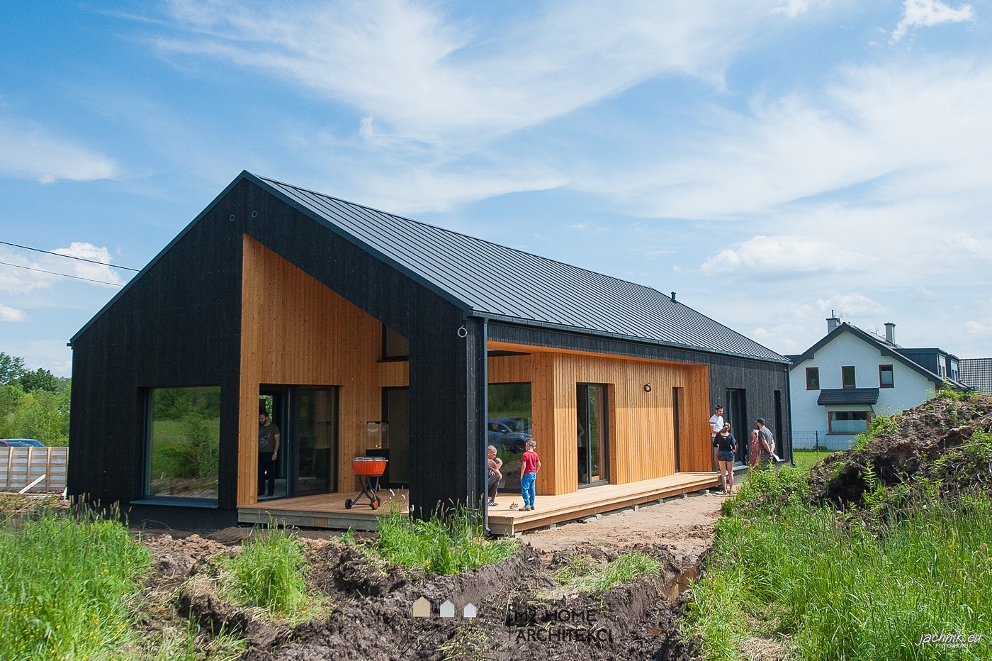 Nowoczesny dom szkieletowy z czarną elewacją i drewnianymi panelami, z tarasem i grillem, częściowo zagospodarowany teren wokół domu, widoczni ludzie.