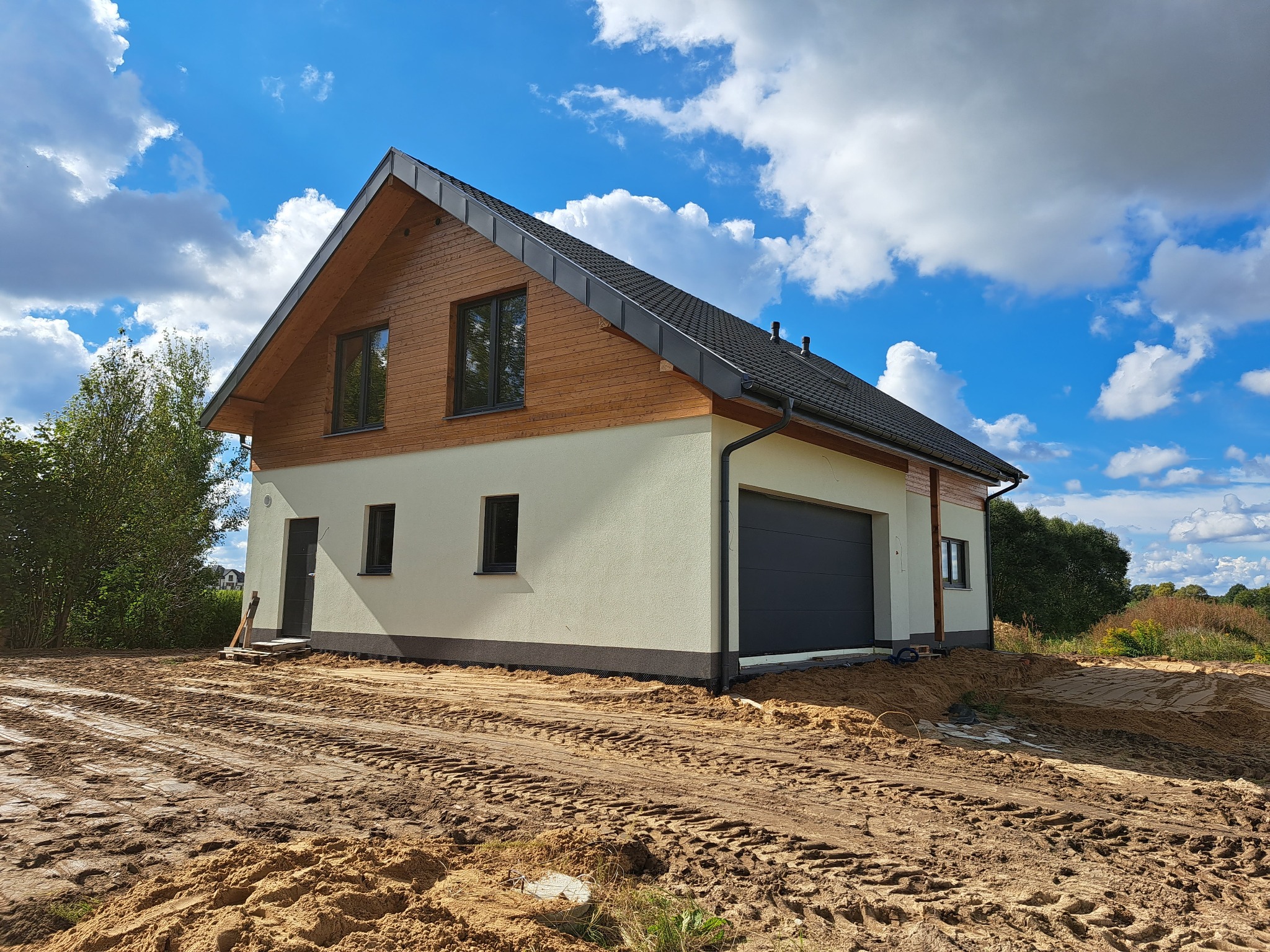 Nowoczesny dom z drewnianą elewacją na poddaszu, grafitowa brama garażowa, teren wokół domu w trakcie prac ziemnych, błękitne niebo z białymi chmurami.
