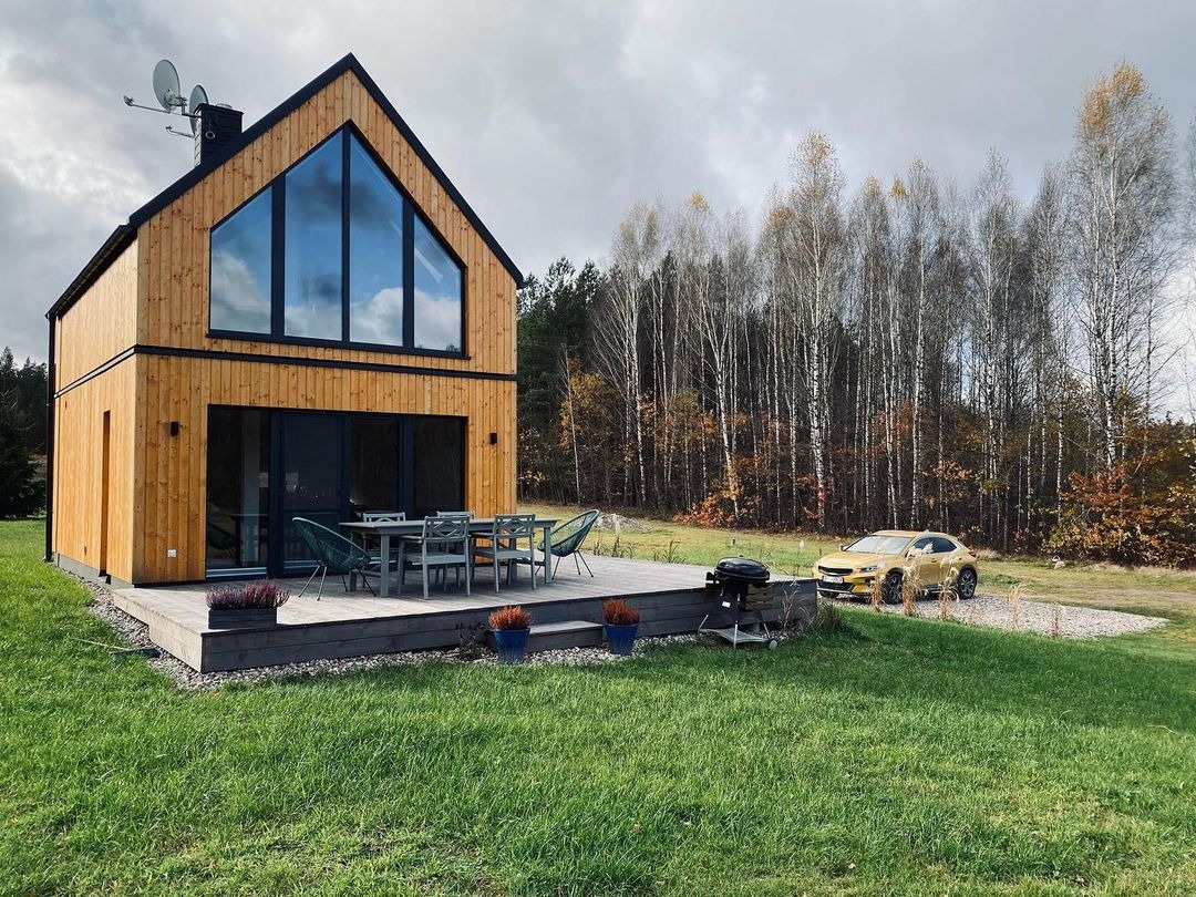 Dwukondygnacyjny dom z drewna z dużym trójkątnym oknem, tarasem z meblami ogrodowymi i żółtym samochodem na tle lasu.