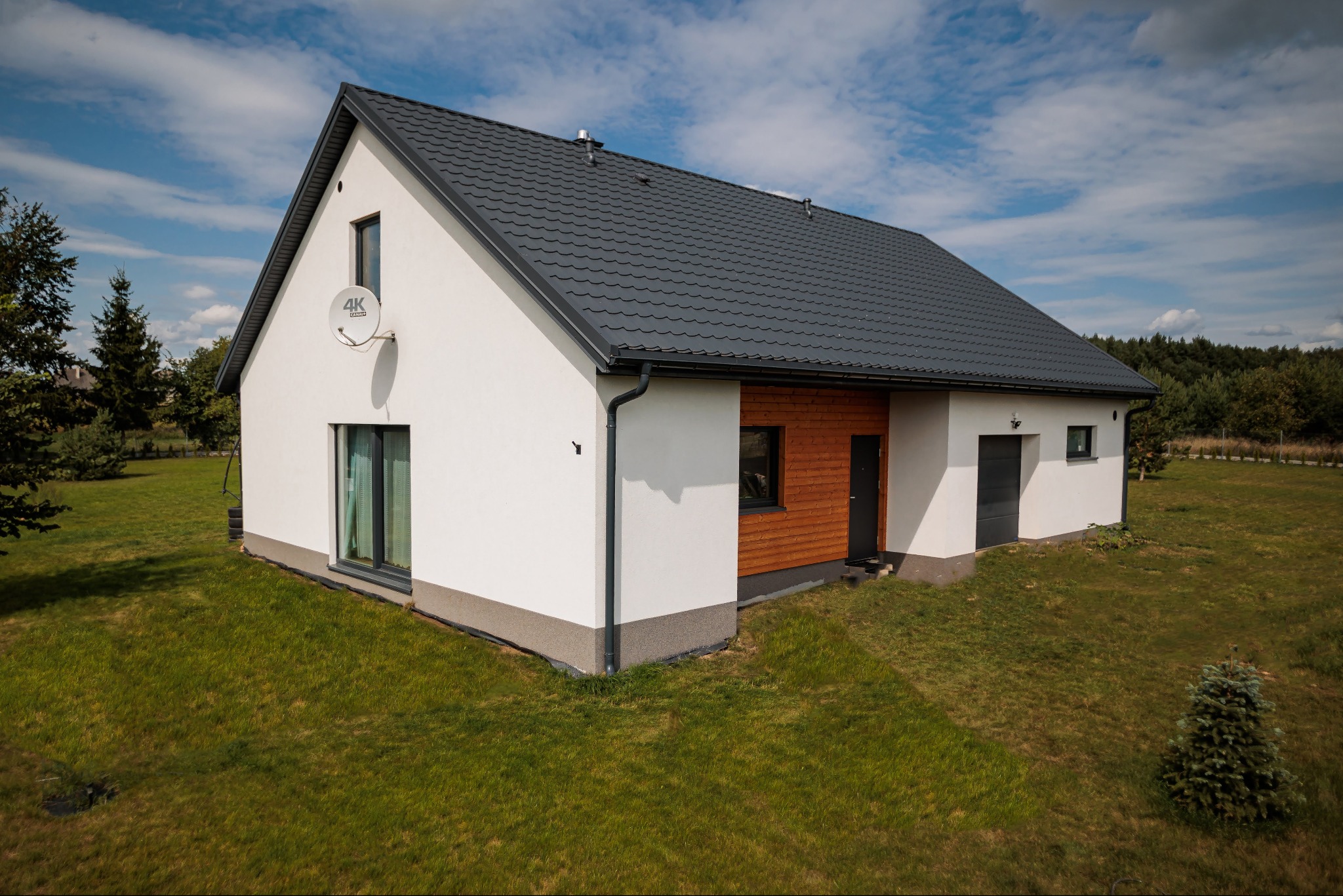 Nowoczesny dom jednorodzinny z białymi ścianami, grafitowym dachem i drewnianą elewacją, otoczony zielonym trawnikiem i drzewami, z anteną satelitarną na ścianie.