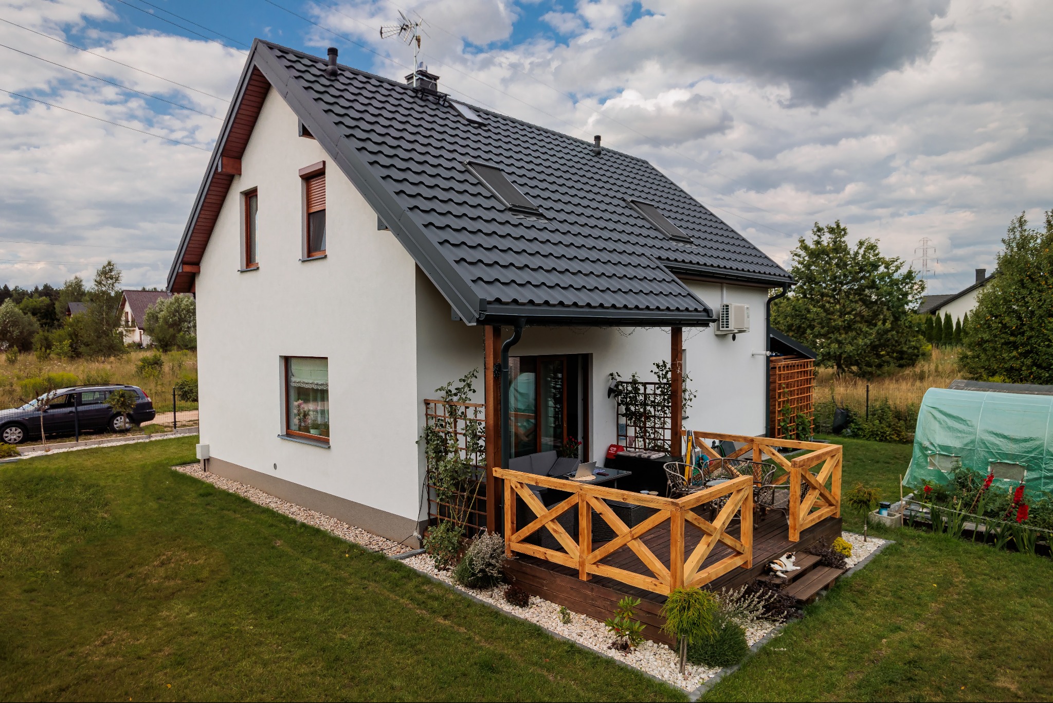 Nowoczesny, biały dom z ciemną dachówką, drewnianym tarasem i zadbanym ogrodem w słoneczny dzień.
