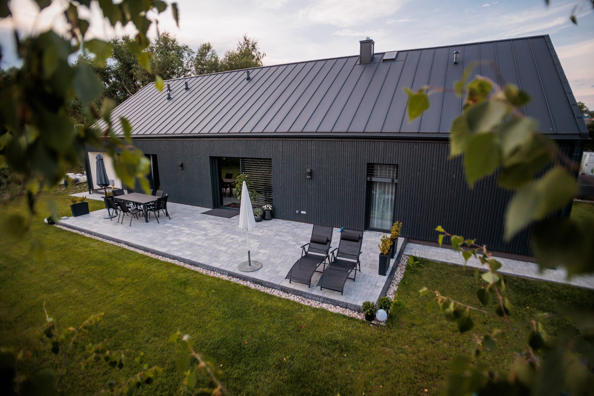 Nowoczesny dom szkieletowy z ciemną elewacją i metalowym dachem, widok z góry na taras z meblami ogrodowymi i zadbanym trawnikiem, liście drzew na pierwszym planie