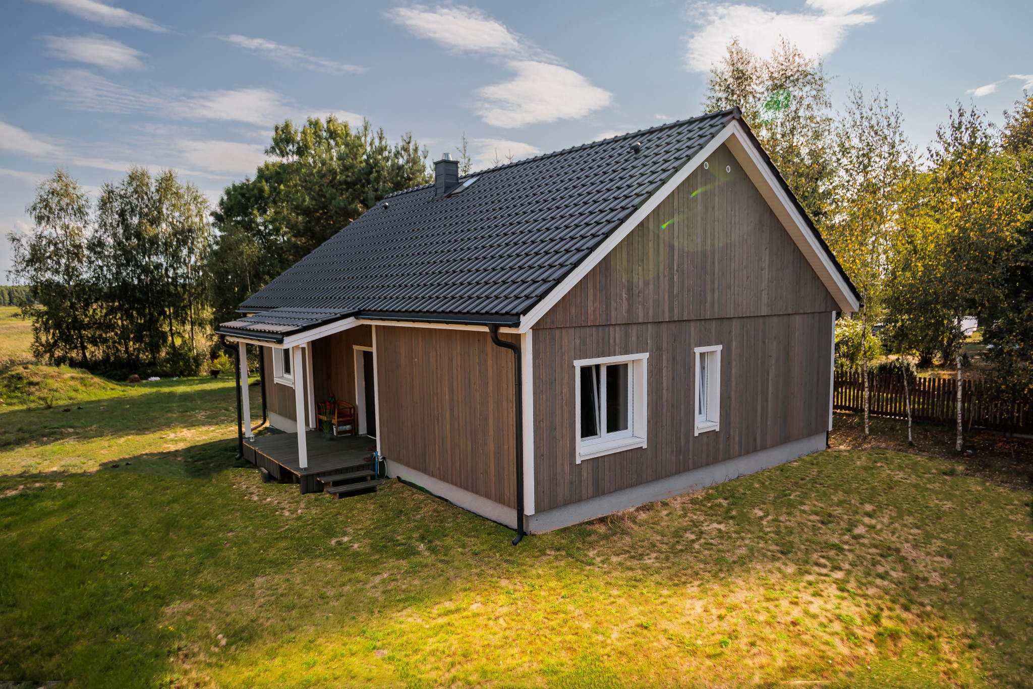 Gotowy, parterowy budynek z drewnianą elewacją i ciemną dachówką, z małym zadaszonym tarasem, otoczony zielonym trawnikiem i drzewami.