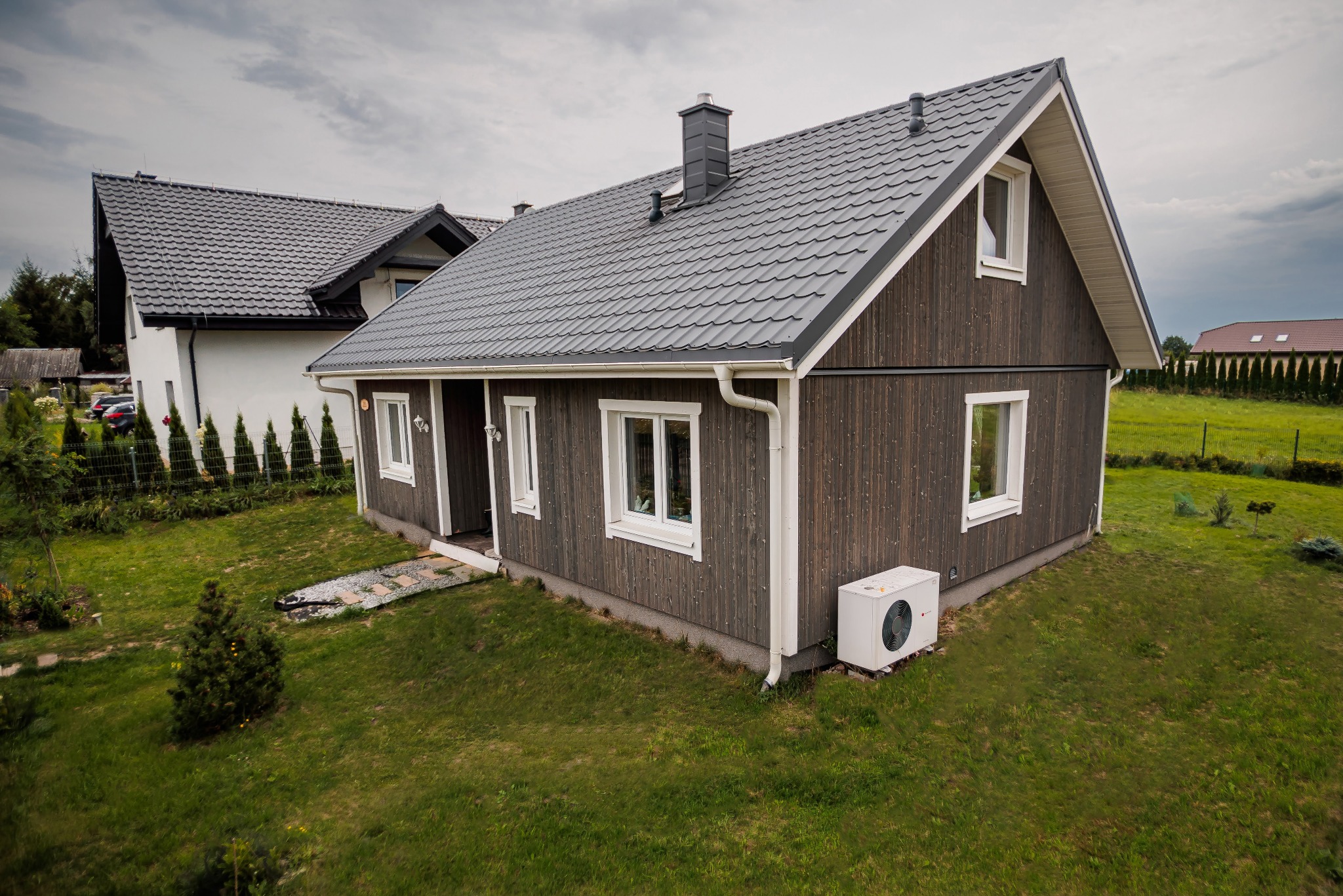 Nowoczesny dom z ciemnego drewna z szarą dachówką, widok z góry na tle zielonego trawnika i pochmurnego nieba, z widoczną jednostką klimatyzacji zewnętrznej przy ścianie.
