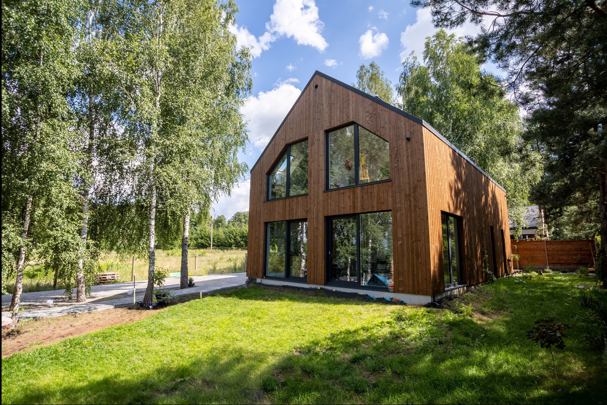 Nowoczesny dom z drewnianą elewacją i dużymi oknami, otoczony zielonym trawnikiem i drzewami brzozy w słoneczny dzień.
