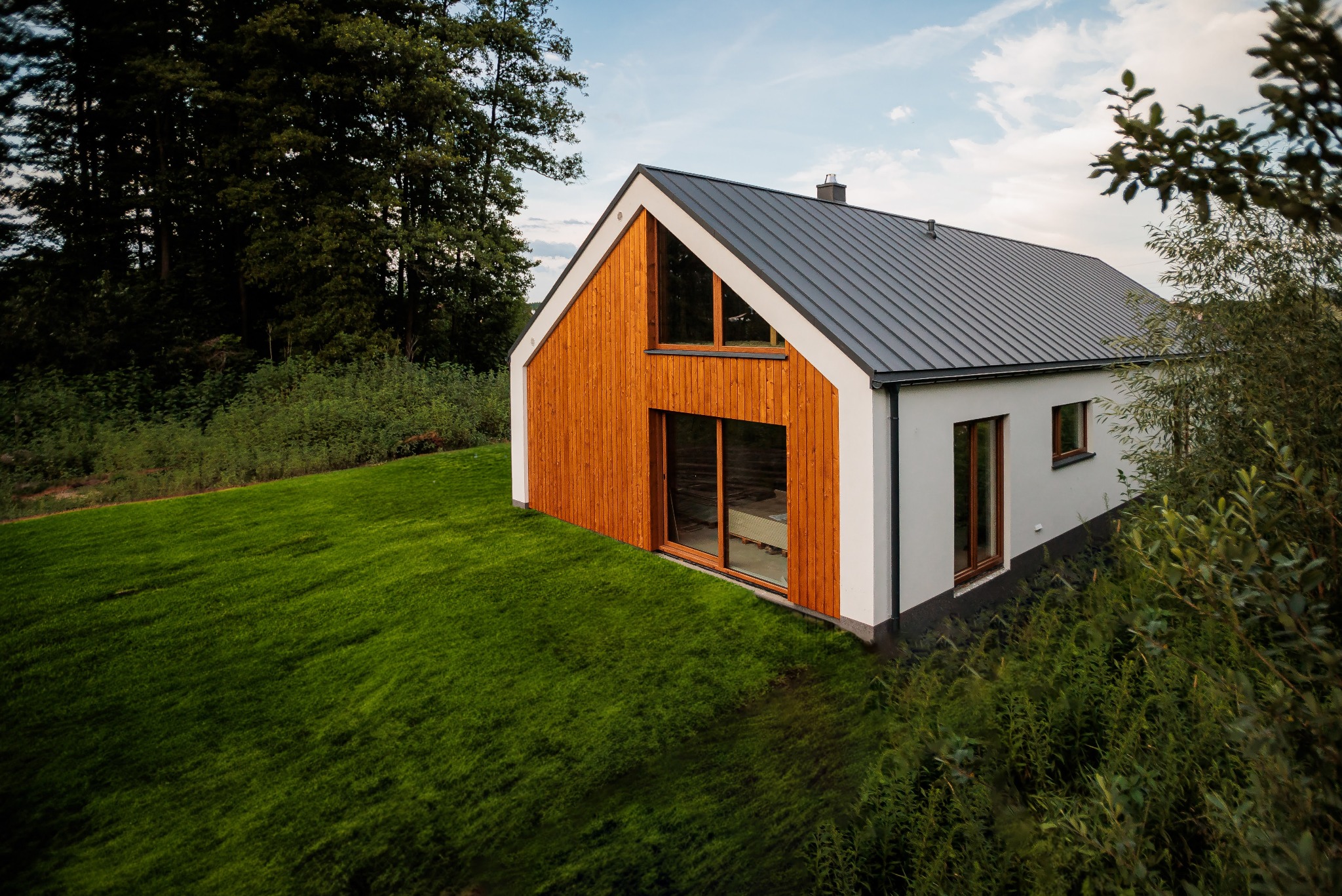 Nowoczesny dom z drewnianą elewacją i szarą blachodachówką, otoczony zielonym trawnikiem i drzewami, widziany z perspektywy lekko z góry.