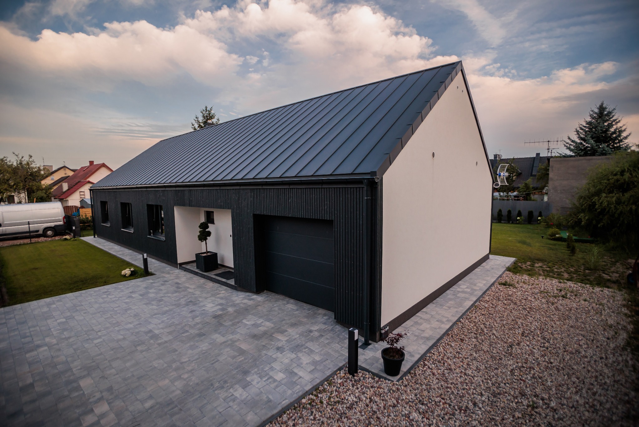 Nowoczesny dom z grafitową blachą na dachu, elewacją z pionowych desek i jasną ścianą szczytową, widok z góry na brukowany podjazd.