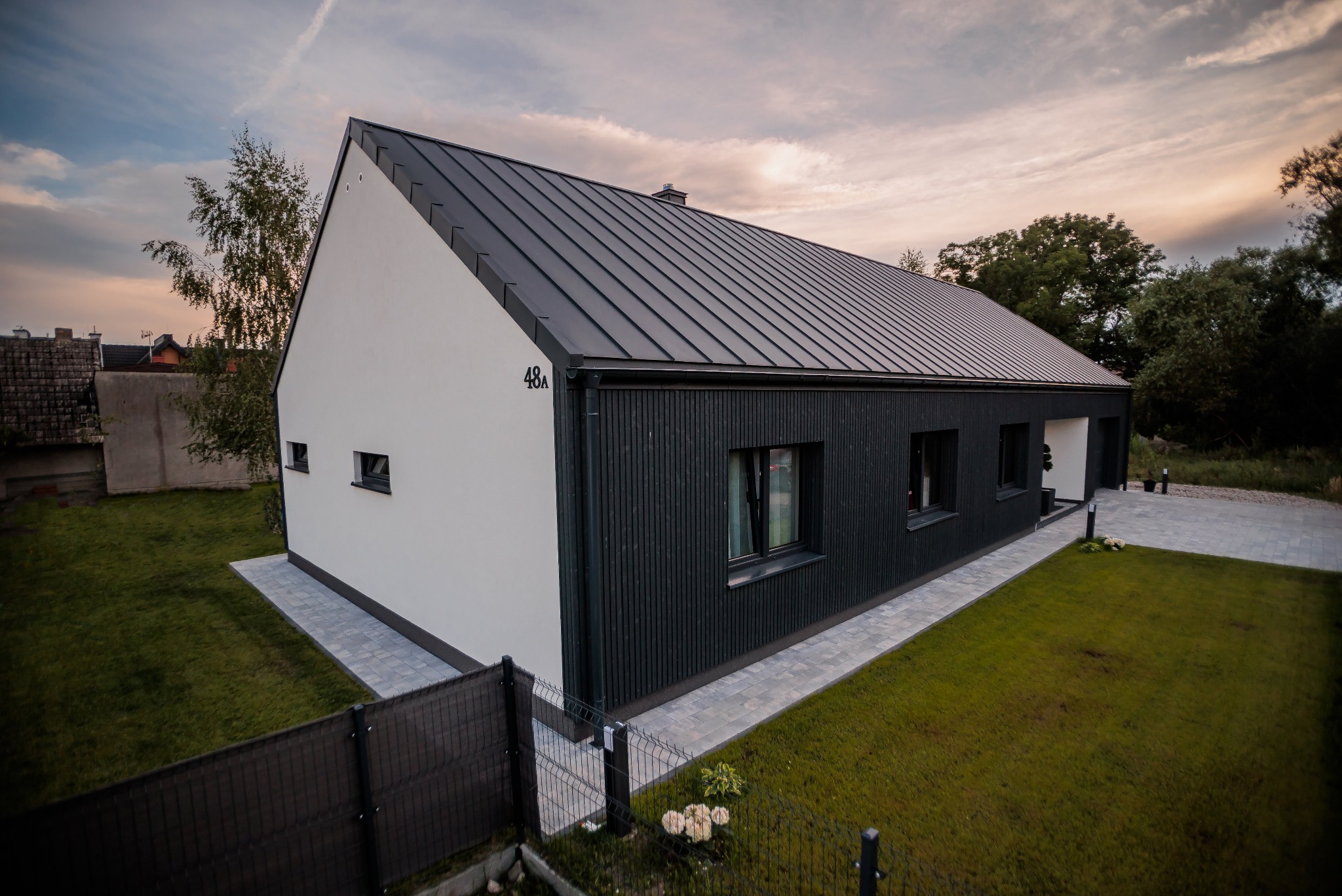 Nowoczesny dom z białą fasadą i ciemną, metalową blachą na dachu, numer domu 48A, otoczony trawnikiem i ogrodzeniem z siatki.