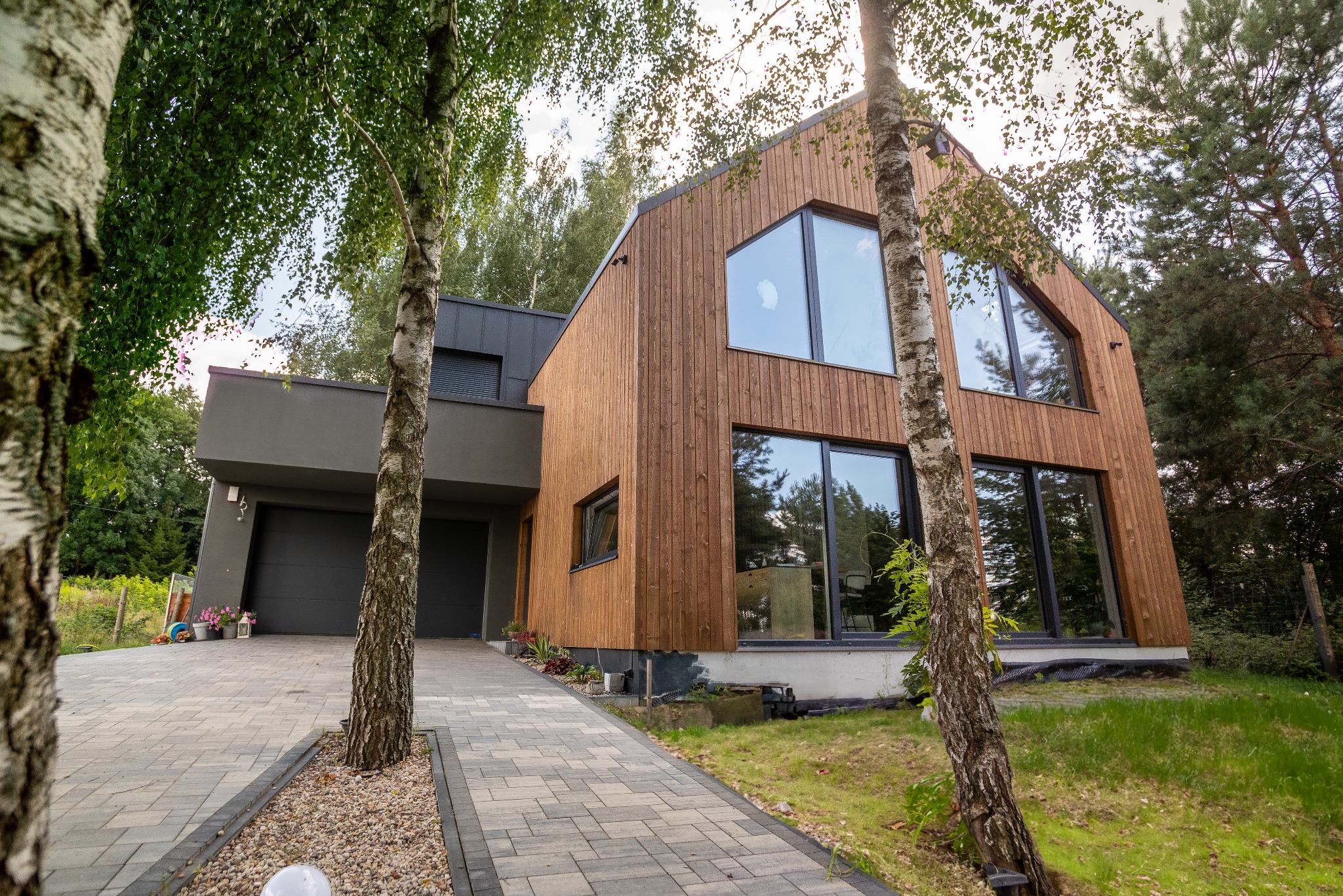Nowoczesny dom szkieletowy z drewnianą elewacją i dużymi oknami, widoczny z perspektywy chodnika z kostki brukowej, otoczony drzewami.