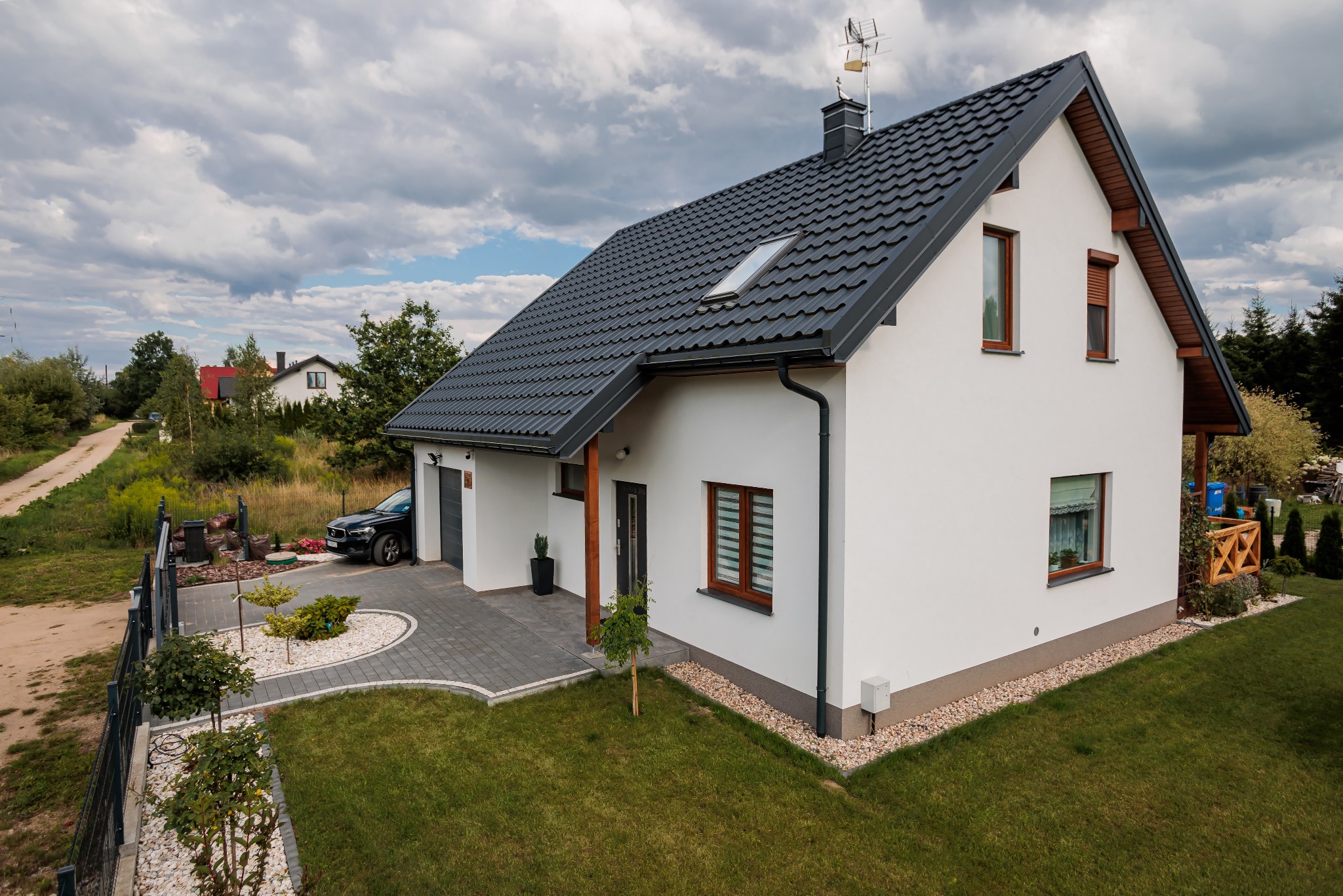 Nowoczesny, biały dom z ciemną, skośną dachówką, widok z góry na ogród z kostką brukową i zaparkowanym samochodem, otoczony zielenią i innymi domami w oddali.