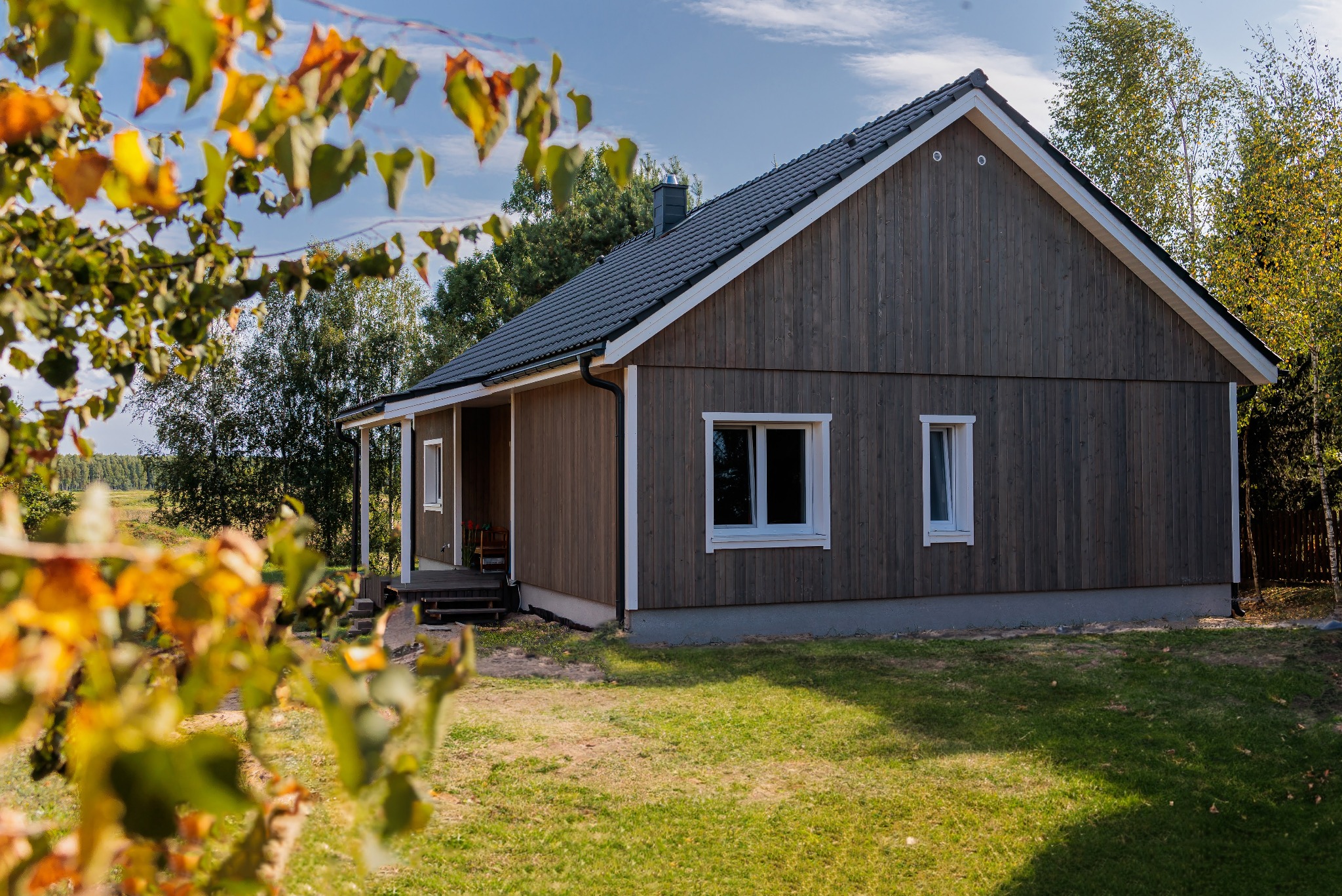 Gotowy, parterowy dom z ciemnego drewna z białymi ramami okiennymi, widoczny zza drzew z jesiennymi liśćmi, na zadbanym trawniku.
