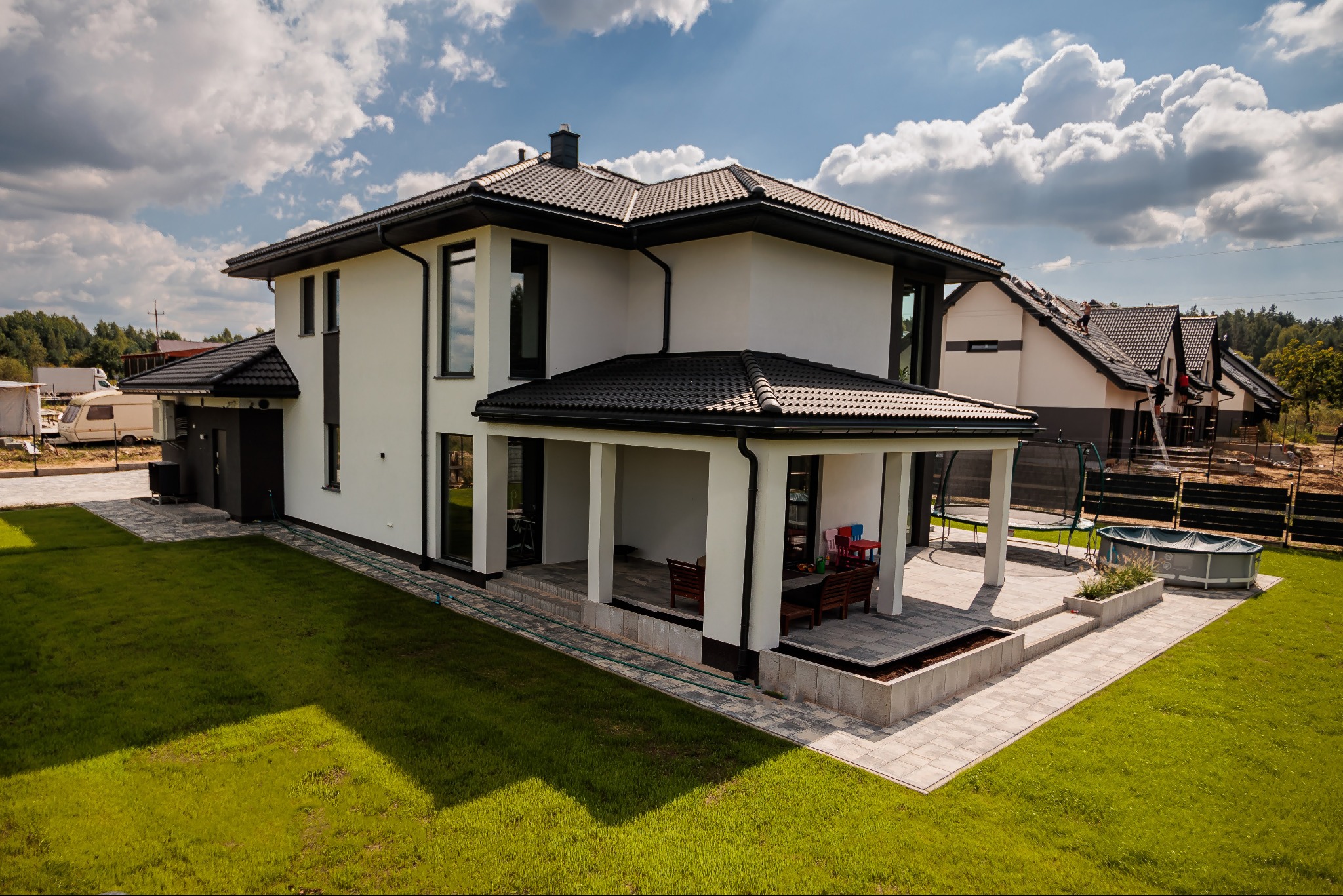 Nowoczesny dom jednorodzinny z białymi ścianami i ciemną dachówką, widok z lotu ptaka na ogród z trampoliną i basenem.