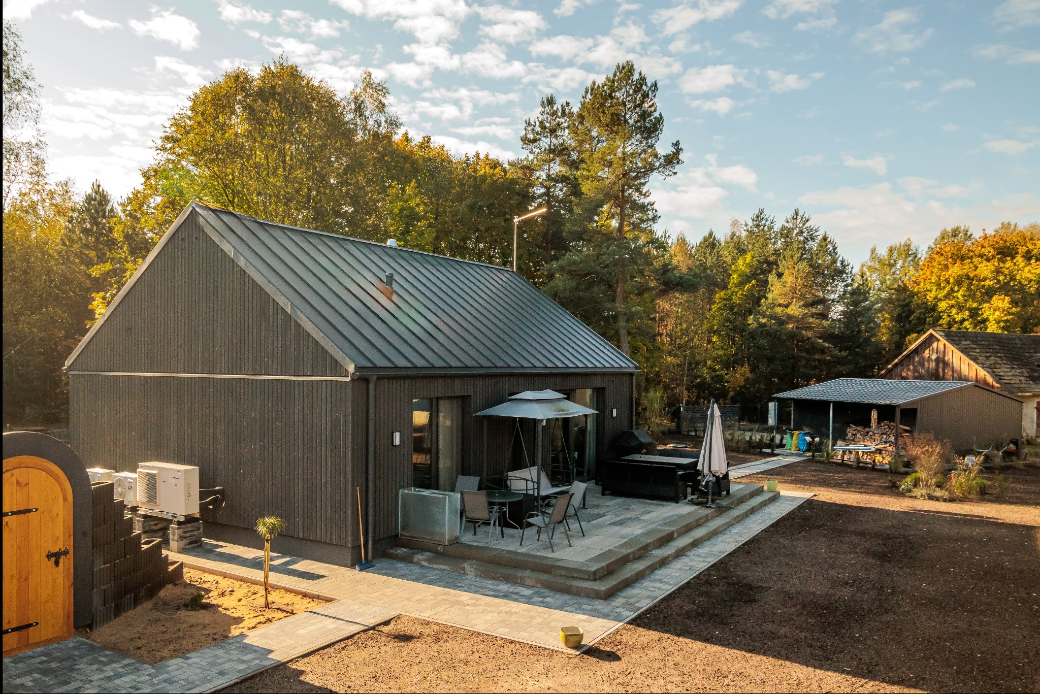 Nowoczesny, parterowy dom z ciemnego drewna z metalowym dachem, tarasem i otoczeniem z drzew w jesiennej scenerii.