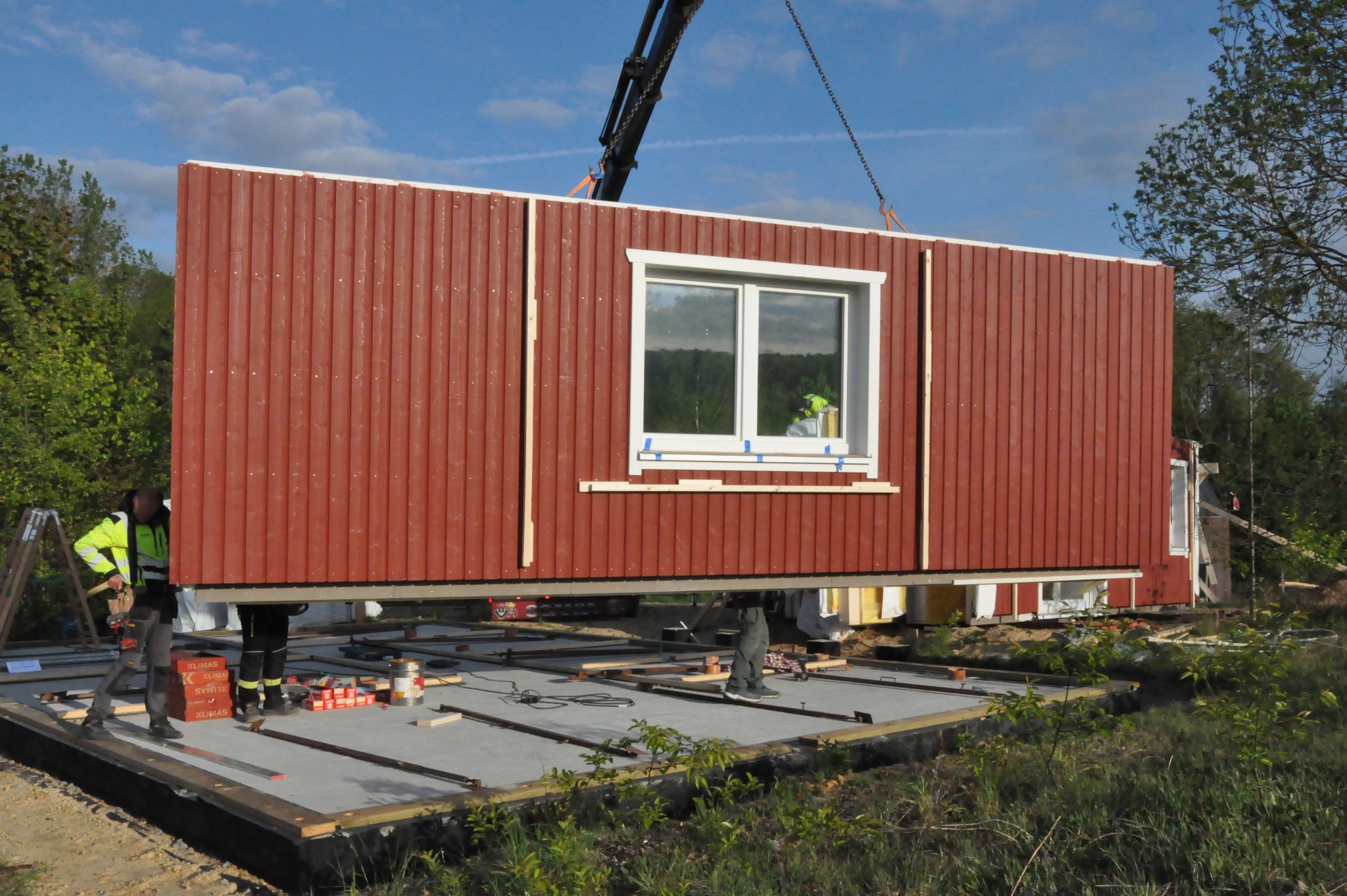 Montaż ściany z oknem na fundamencie domu modułowego za pomocą dźwigu. Widoczni pracownicy i narzędzia. Ściana w kolorze czerwonym, na tle zieleni i błękitnego nieba.
