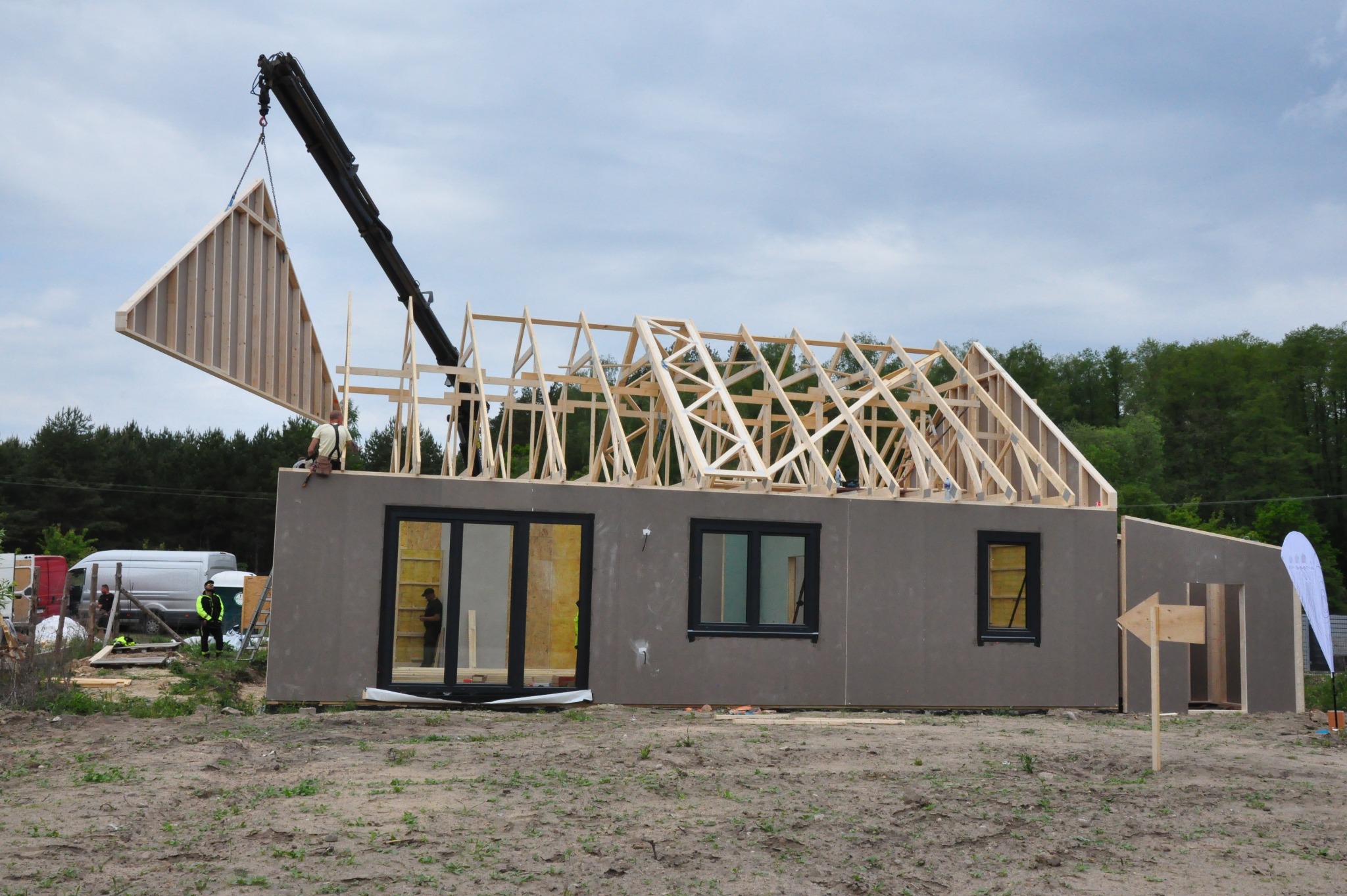 Montaż domu szkieletowego: dźwig podnosi drewniany element konstrukcyjny dachu. Widoczne ściany z oknami i częściowo ukończona więźba dachowa. Etap budowy.