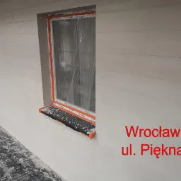 Świeżo otynkowana elewacja budynku z dwoma oknami, zabezpieczonymi folią, widoczne ślady zaprawy na parapecie i wokół okien, faktura tynku imitująca drewniane deski.