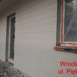 Świeżo wykończona elewacja budynku z jasnymi panelami imitującymi drewno, okna zabezpieczone folią, widoczne ślady zaprawy i gruzu na ziemi, narożnik budynku z widocznymi betonowymi elementami.