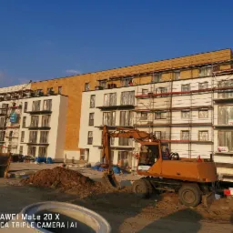 Budynek wielorodzinny w trakcie ocieplania, z widocznymi rusztowaniami i koparką na pierwszym planie, przy słonecznej pogodzie.