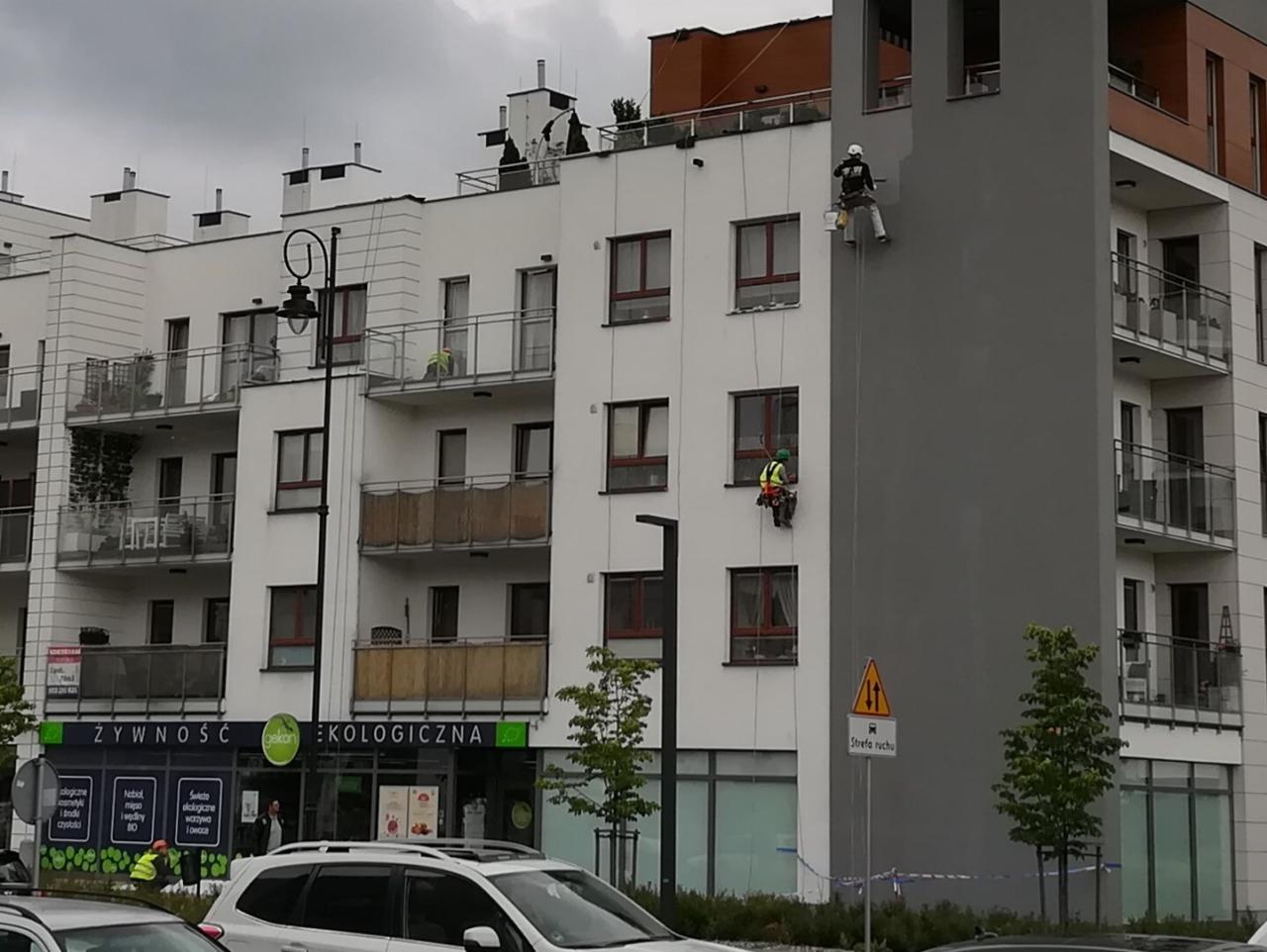 Dwóch alpinistów na linach maluje elewację wysokiego bloku mieszkalnego na szaro, widoczny znak ostrzegawczy strefy ruchu.
