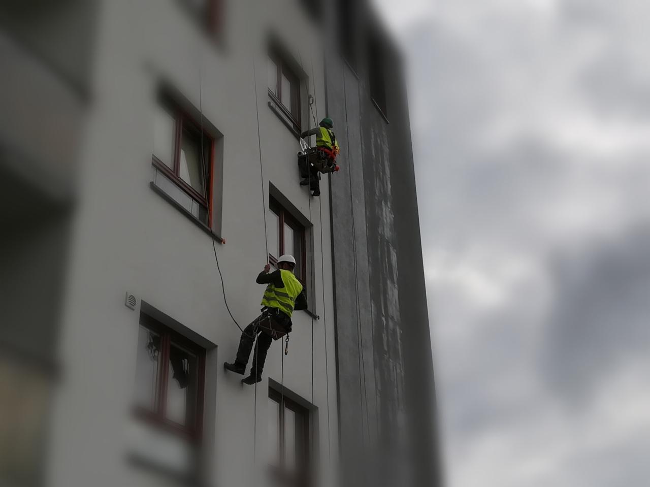Dwóch alpinistów przemysłowych na linach czyści elewację wysokiego budynku z szarego osadu, jeden z nich trzyma w ręku białą szpachelkę.