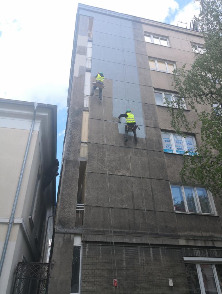 Pracownicy na linach zabezpieczających podczas przygotowywania elewacji budynku we Wrocławiu do malowania, fragment ściany już zagruntowany.