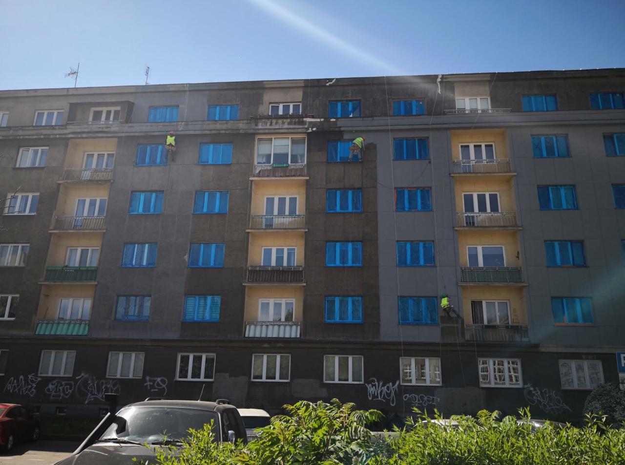 Trzech pracowników na linach myje elewację bloku, część elewacji jest już umyta, okna zabezpieczone folią, widoczne graffiti na dole budynku.
