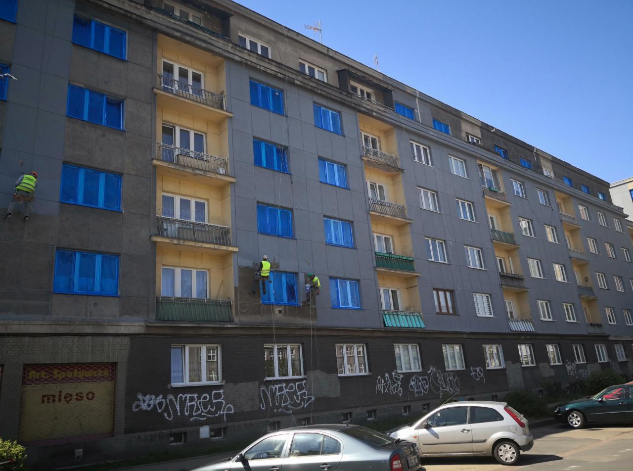 Dwóch pracowników na linach zabezpiecza okna folią przed rozpoczęciem prac na elewacji bloku mieszkalnego, widoczny fragment pomalowanej ściany z graffiti w dolnej części.