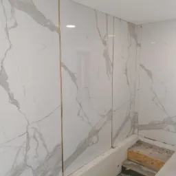 Ściany łazienki wyłożone białymi płytami imitującymi marmur z cienkimi złotymi listwami, widoczny fragment podłogi w trakcie prac wykończeniowych.