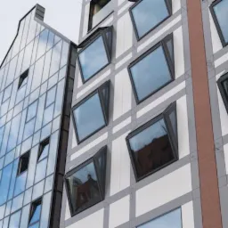 Nowoczesny budynek z oknami aluminiowymi, niektóre z nich uchylone, widok z dołu na elewację i sąsiedni budynek z przeszkloną fasadą.