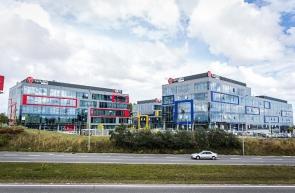 Nowoczesny budynek biurowy z fasadą ze szkła i aluminium, z kolorowymi akcentami w postaci czerwonych, niebieskich i żółtych ram okiennych, widoczny z perspektywy ulicy z samochodem w tle...