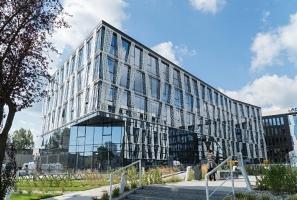 Nowoczesny budynek biurowy z fasadą z szarych paneli i dużymi oknami, widoczny fragment błękitnego nieba z chmurami, metalowe barierki i zieleń przed budynkiem.