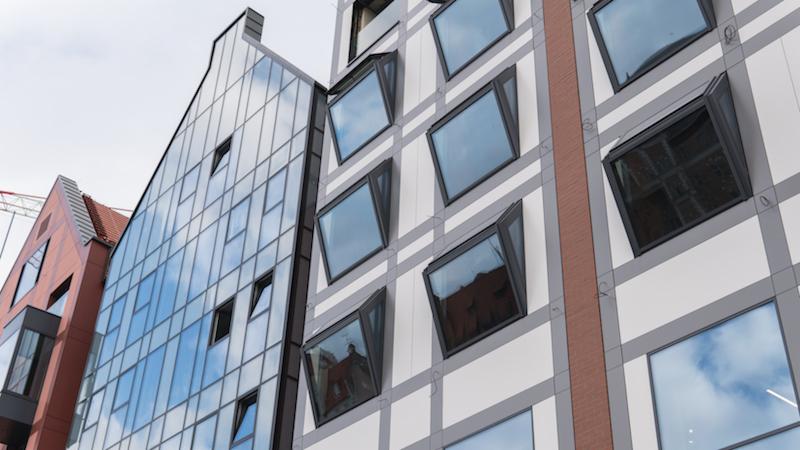Nowoczesny budynek z oknami aluminiowymi, niektóre z nich uchylone, widok z dołu na elewację i sąsiedni budynek z przeszkloną fasadą.