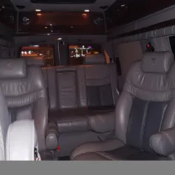 Wnętrze luksusowego busa z szarymi sk&oacute;rzanymi fotelami, widoczne trzy rzędy siedzeń, oświetlenie sufitowe i okna z roletami.