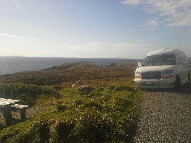 Biały bus zaparkowany na tle malowniczego krajobrazu z oceanem i wzgórzami, obok drewnianego stołu piknikowego.