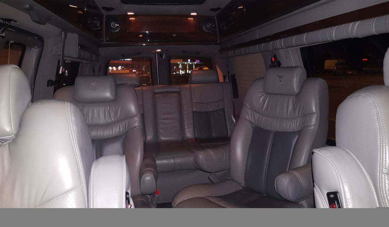Wnętrze luksusowego busa z szarymi skórzanymi fotelami, widoczne trzy rzędy siedzeń, oświetlenie sufitowe i okna z roletami.