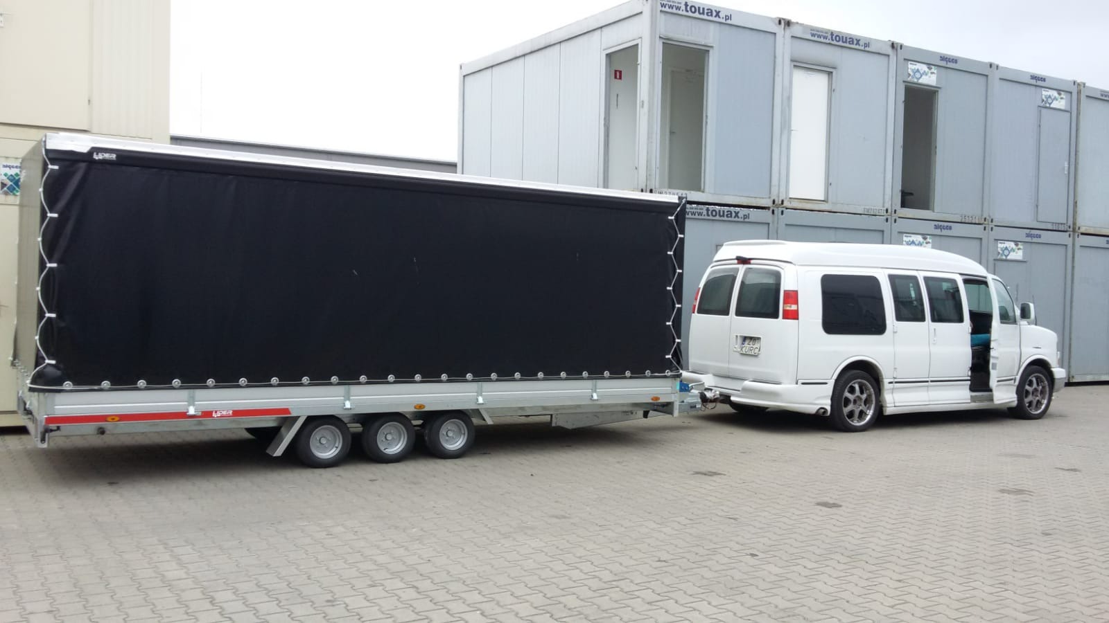 Biały van z otwartymi drzwiami holuje przyczepę z czarną plandeką na tle kontenerów biurowych Touax.