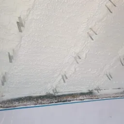 Świeżo nałożona, biała izolacja piankowa z widocznymi metalowymi mocowaniami na skośnym poddaszu, zabezpieczona folią ochronną.