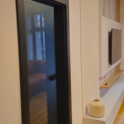 Energo-Tech Paweł Warszewski - Nowoczesne drzwi wewnętrzne z czarną ramą i szklanym wypełnieniem, odbijające wnętrze salonu z beżową sofą i drewnianą podłogą. Minimalistyczny design.