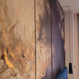 Energo-Tech Paweł Warszewski - Panel ścienny z abstrakcyjnym wzorem w odcieniach złota i szarości, zamontowany za wezgłowiem łóżka z niebieską tapicerką. Wiszące lampy po bokach.