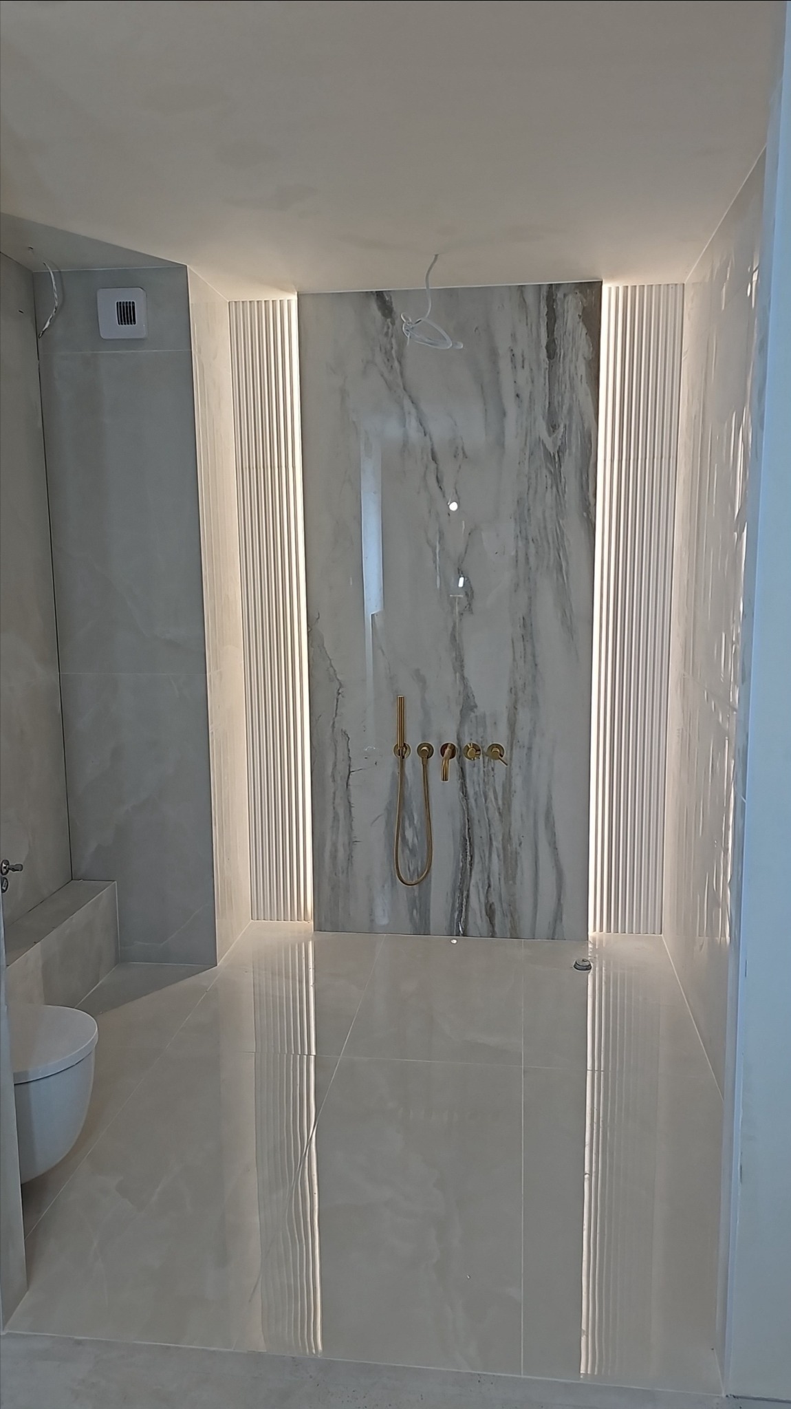 Wykończona kabina prysznicowa z marmurową ścianą i złotą armaturą w Poznaniu. Podświetlane panele dekoracyjne po bokach. Błyszczące płytki podłogowe.