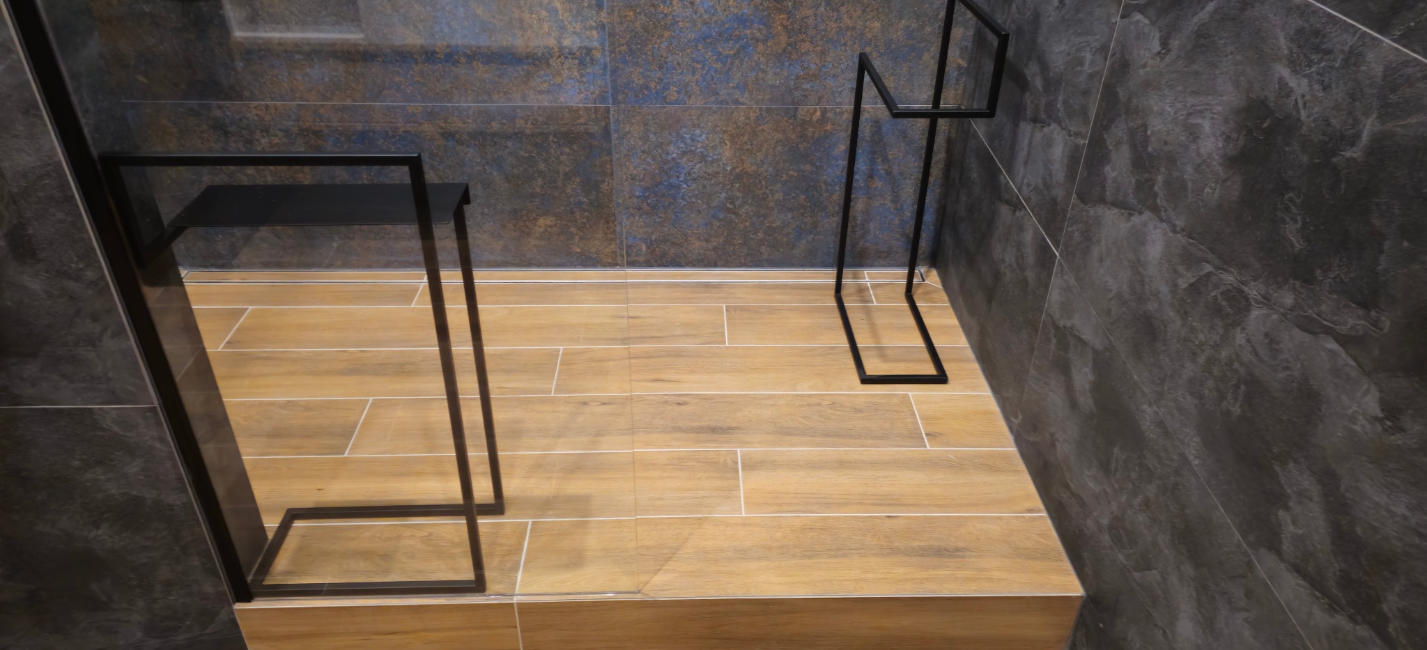 Ułożone panele podłogowe imitujące drewno w łazience z ciemnymi, marmurkowymi płytkami ściennymi i minimalistycznymi, czarnymi akcesoriami łazienkowymi.