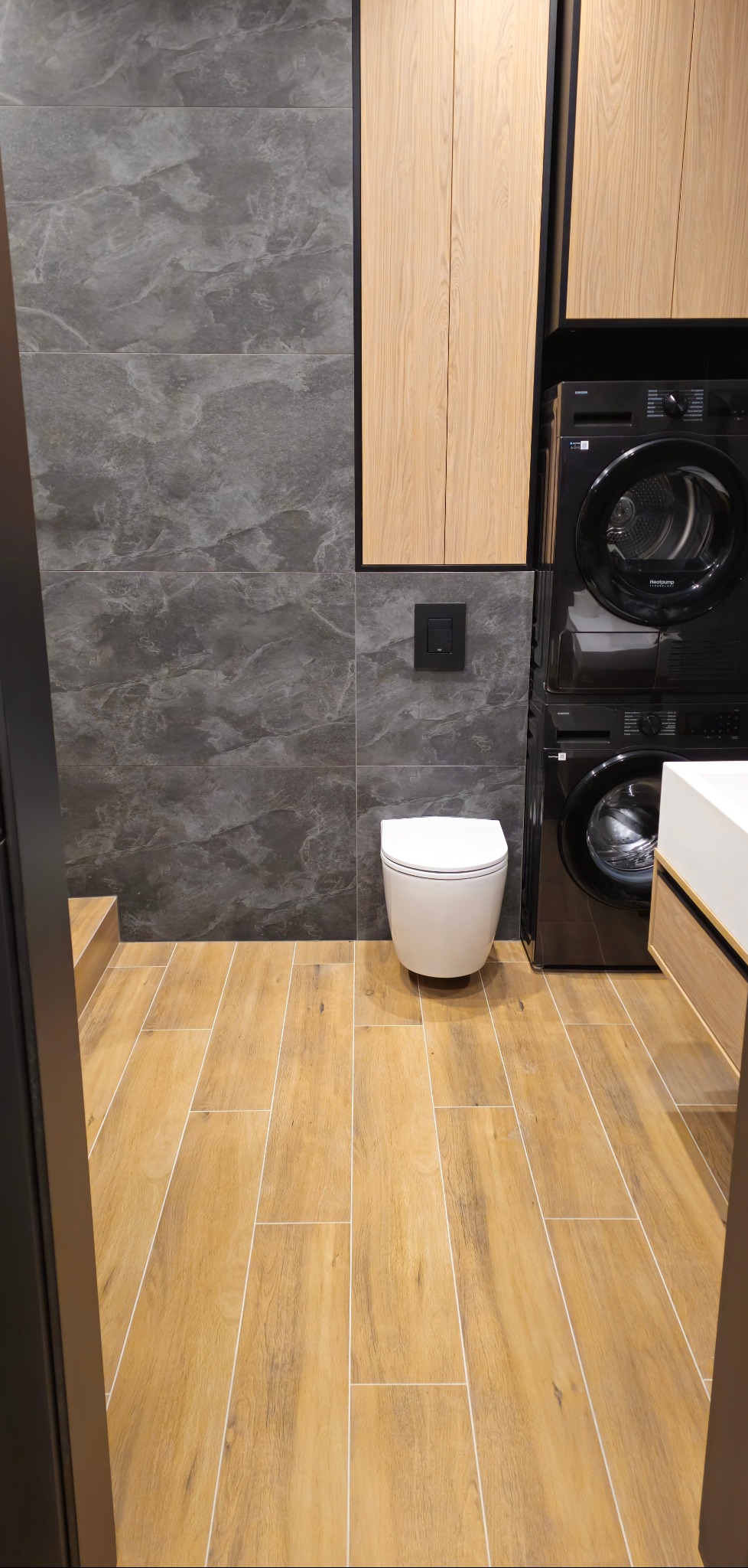 Nowoczesna łazienka z drewnopodobną podłogą, szarą ścianą z płytek, podwieszaną toaletą i pralko-suszarką w zabudowie. Minimalistyczny design, ciepłe i chłodne barwy.