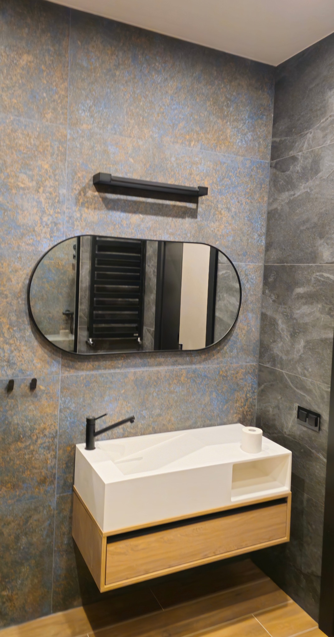 Nowoczesna łazienka z owalnym lustrem, białą umywalką na drewnianej szafce i grafitowymi płytkami na ścianach. Widoczny czarny kran i grzejnik drabinkowy w odbiciu lustra.