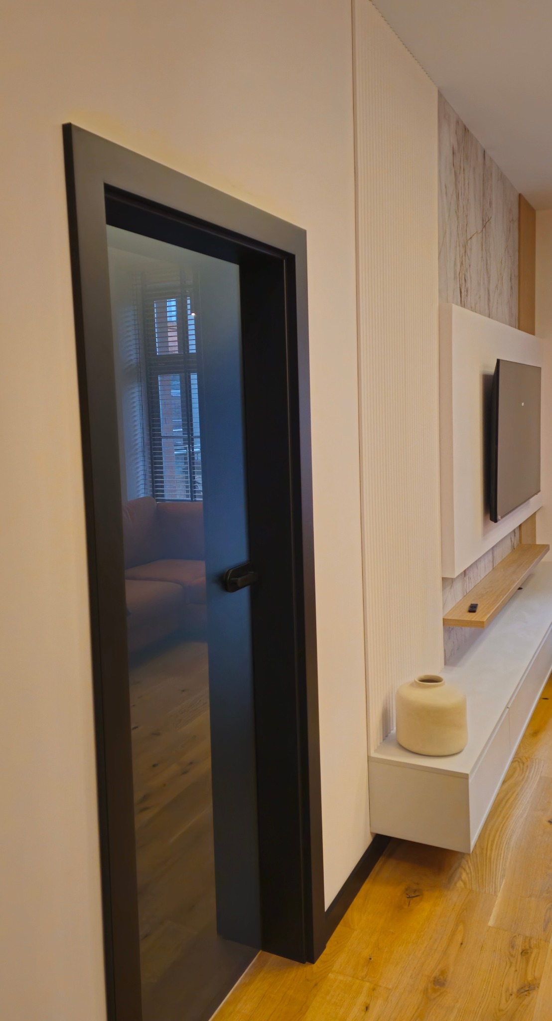 Nowoczesne drzwi wewnętrzne z czarną ramą i szklanym wypełnieniem, odbijające wnętrze salonu z beżową sofą i drewnianą podłogą. Minimalistyczny design.