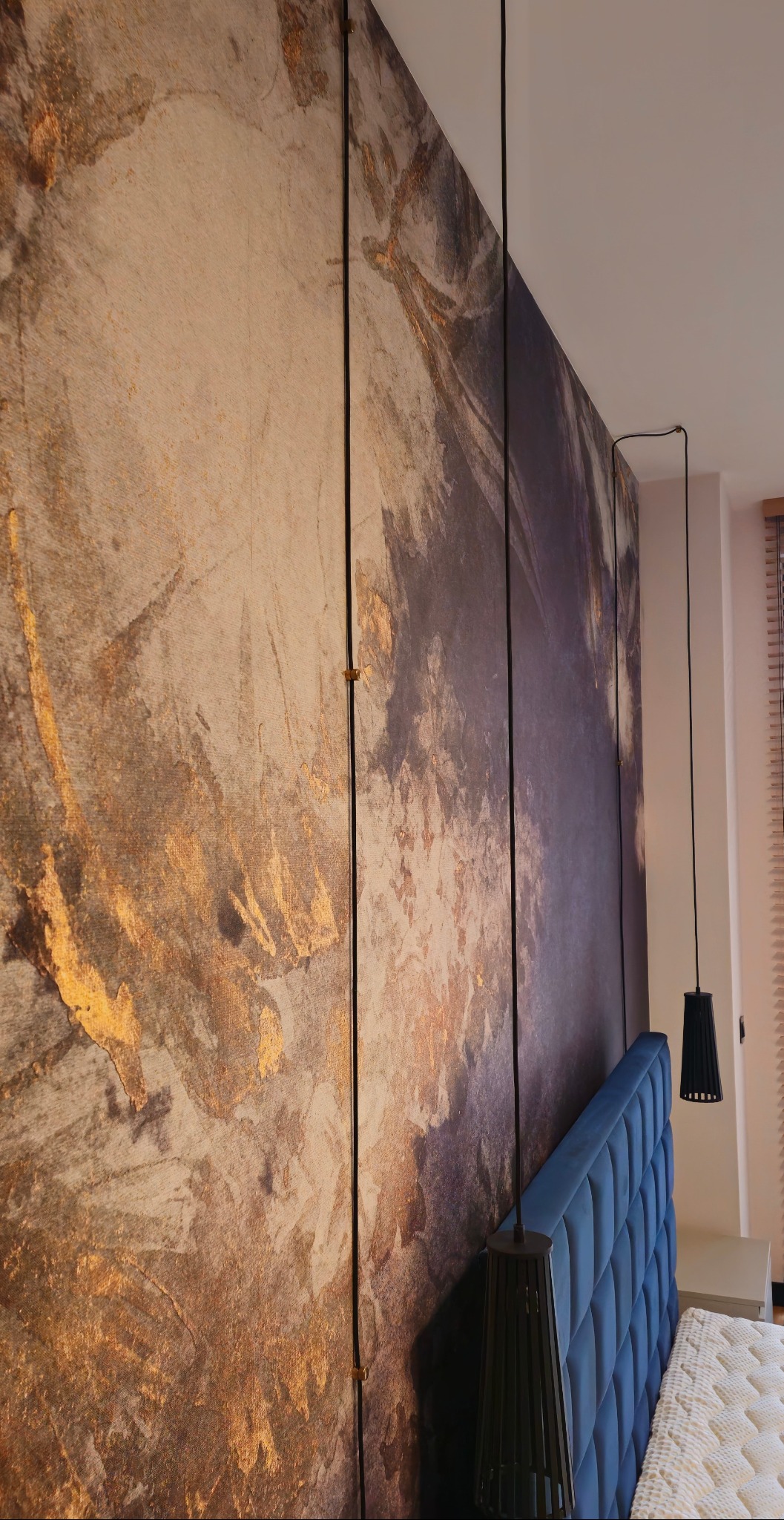Panel ścienny z abstrakcyjnym wzorem w odcieniach złota i szarości, zamontowany za wezgłowiem łóżka z niebieską tapicerką. Wiszące lampy po bokach.